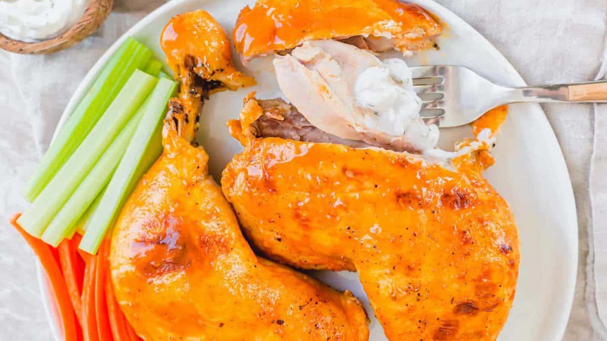 Two pieces of baked chicken with buffalo sauce on a plate, served with celery, carrot sticks, and a side of ranch dressing.