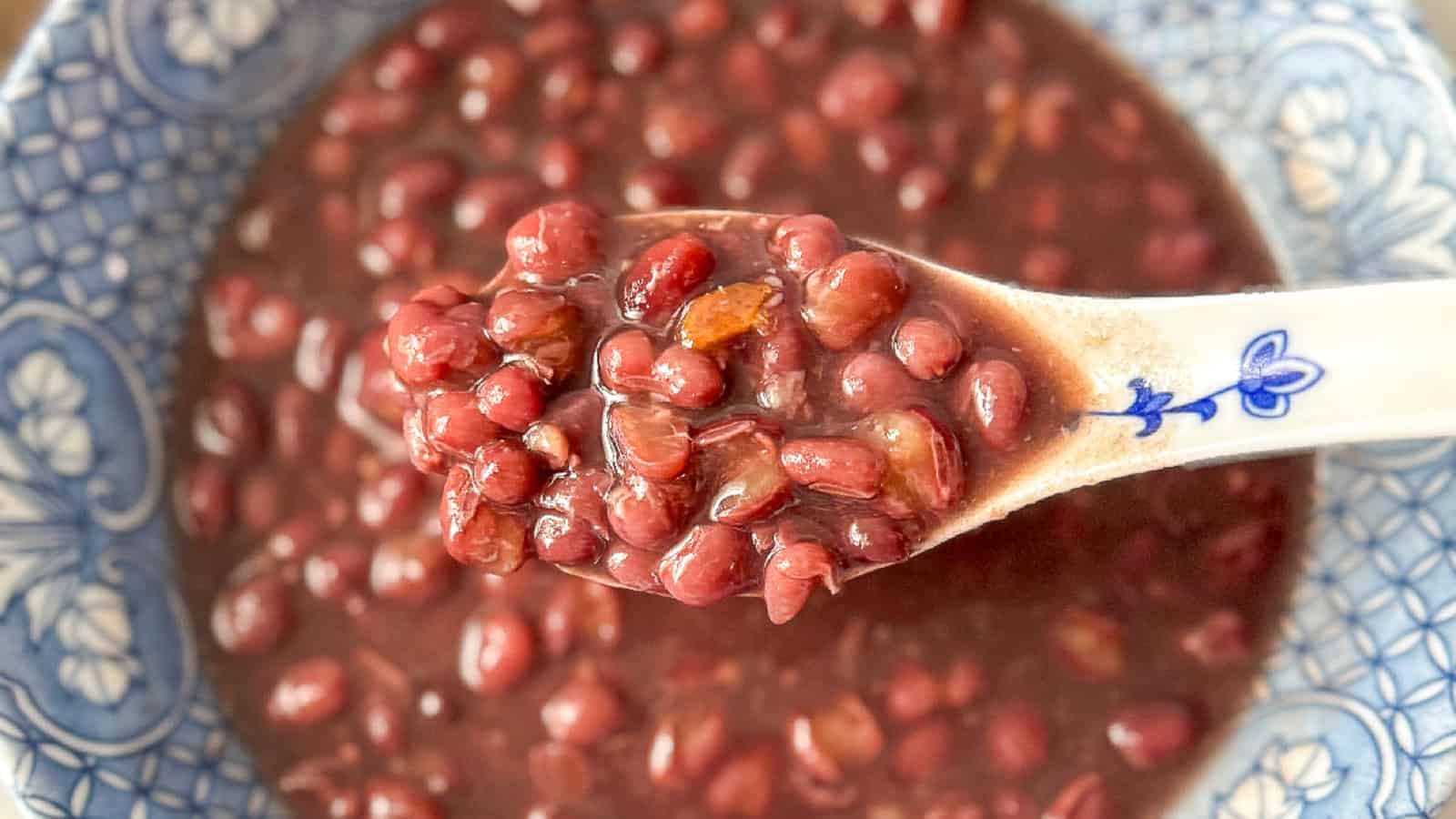 A ceramic spoon holding cooked red beans in a bowl of red bean soup with a blue and white patterned rim.