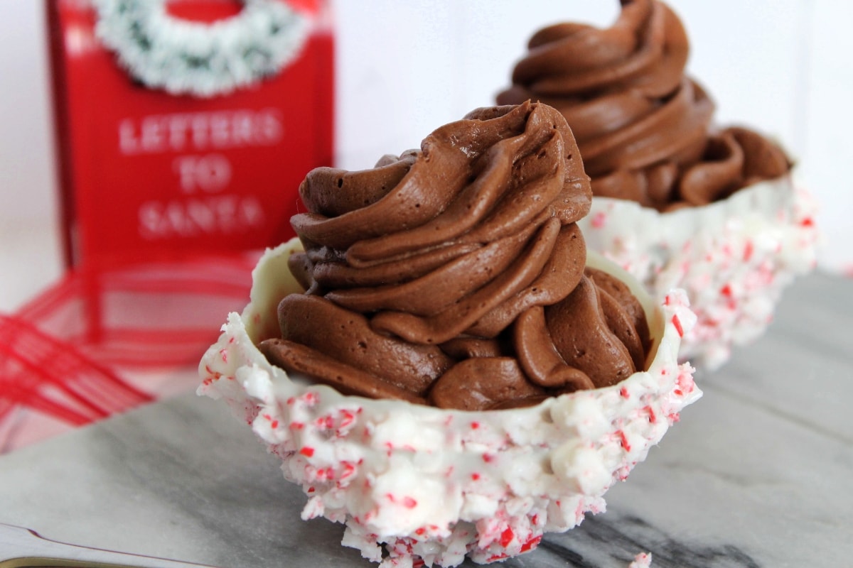Two dessert cups with chocolate mousse in white chocolate bowls coated with crushed peppermint, with a Letters to Santa box in the background.