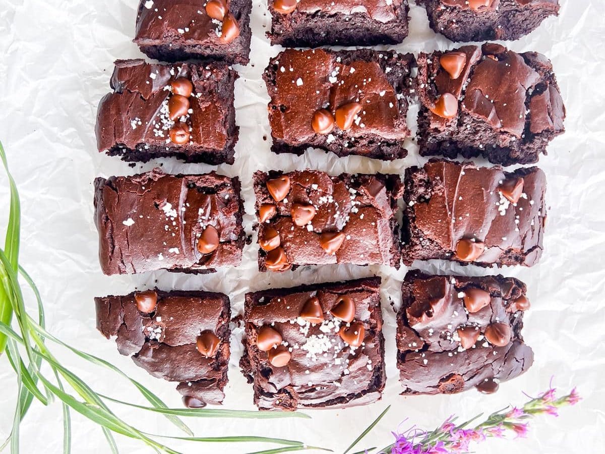 Twelve chocolate brownies topped with chocolate chips and sea salt are arranged on crinkled white parchment paper, with green and purple flowers at the edges.