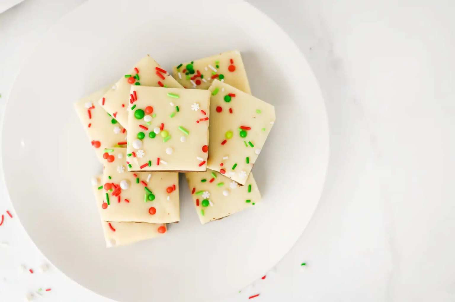 Squares of white chocolate bark topped with red, green, and white sprinkles are arranged on a white plate against a white background.