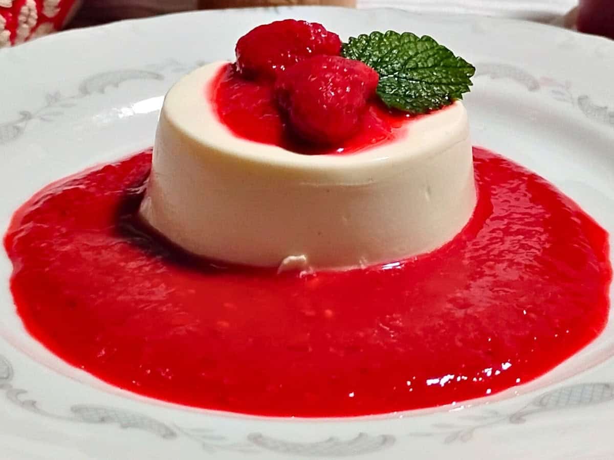 A serving of panna cotta topped with raspberries and a mint leaf, surrounded by raspberry sauce on a white plate.