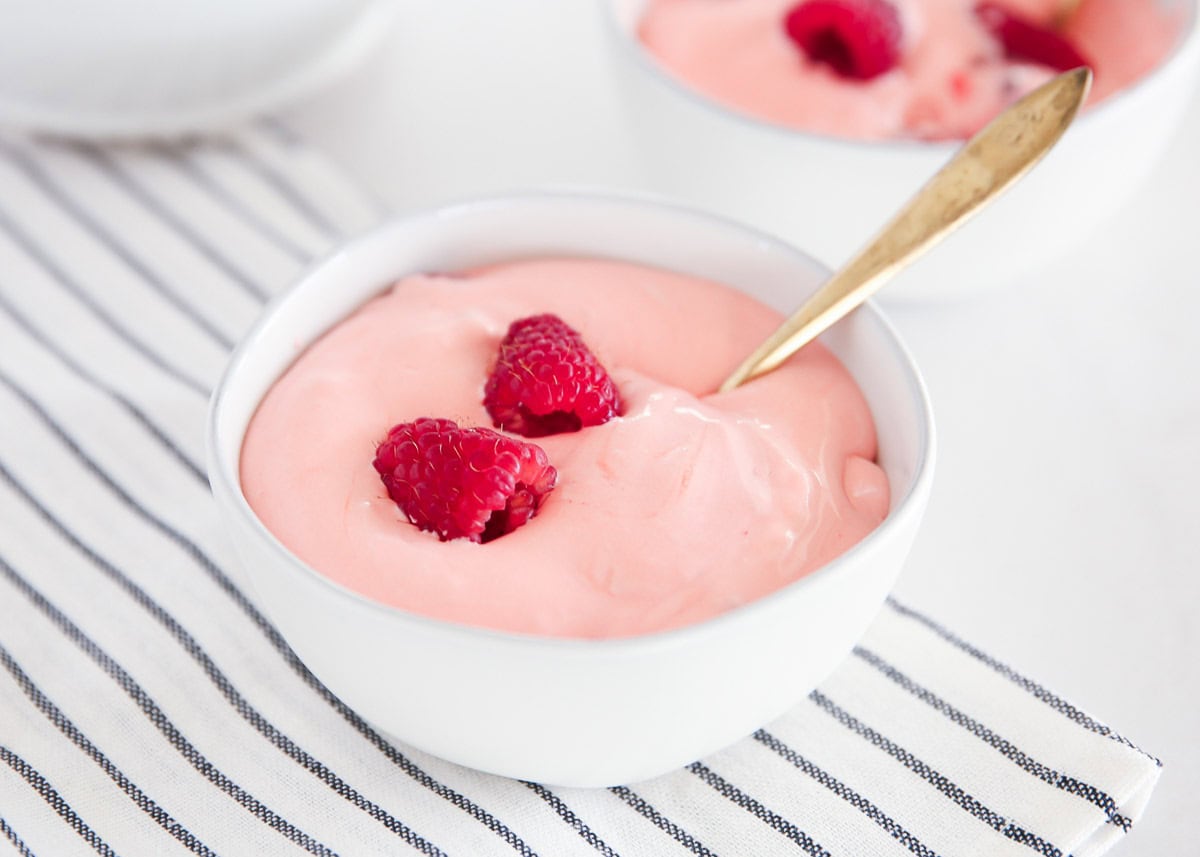 A white bowl filled with pink yogurt topped with two raspberries and a gold spoon, placed on a striped cloth.