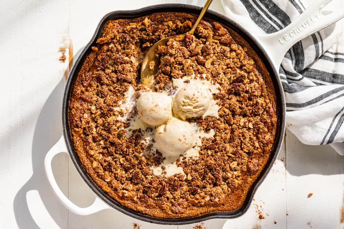 A baked apple crisp topped with three scoops of melting vanilla ice cream in a white skillet, with a serving spoon and striped towel beside it.