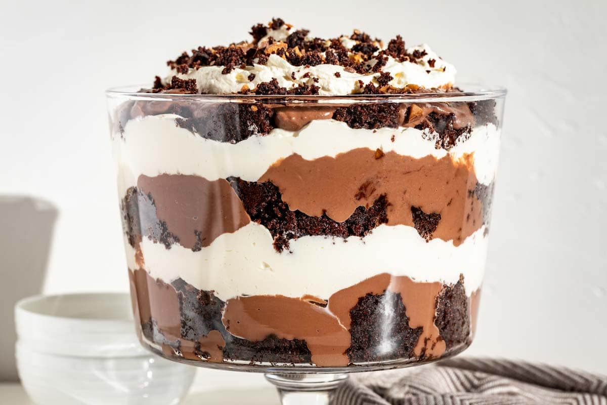 A glass trifle bowl filled with alternating layers of chocolate cake, chocolate pudding, and whipped cream, topped with crumbled cake pieces.
