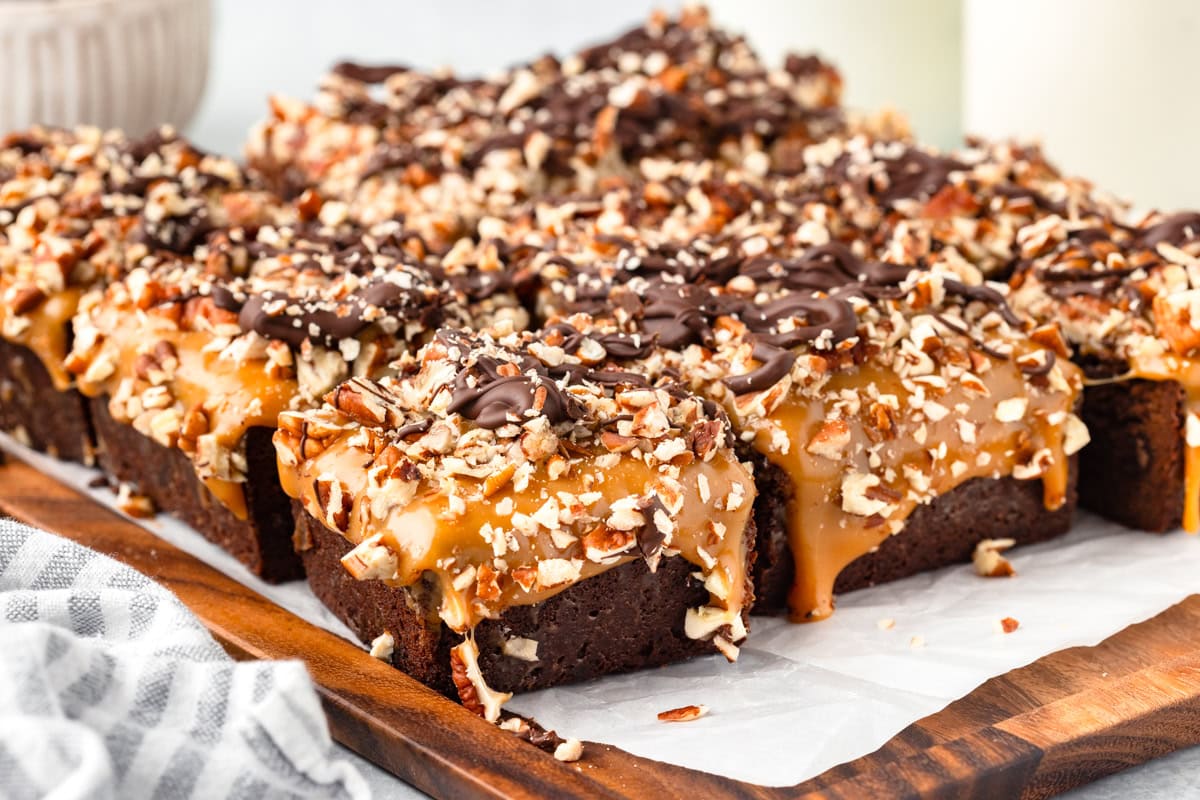 Sliced brownies topped with caramel, chopped pecans, and drizzled chocolate are arranged on a wooden board lined with parchment paper.