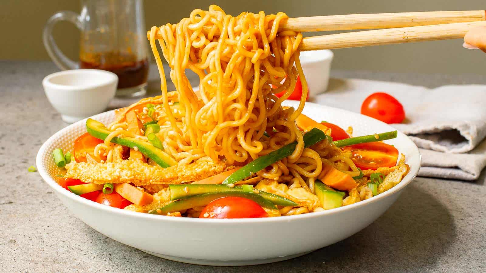 A bowl of noodles with sliced vegetables, including cucumbers and tomatoes, being lifted with chopsticks on a gray countertop.