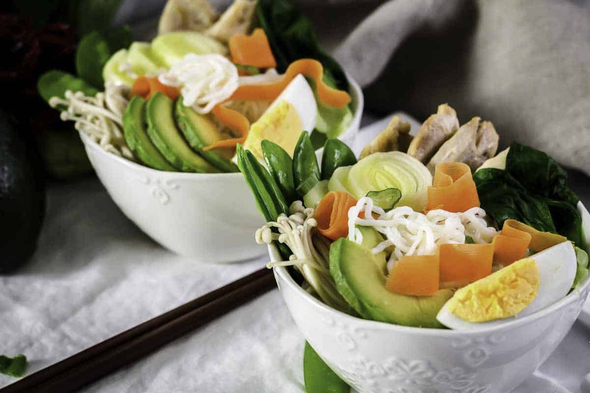 Two bowls of ramen topped with avocado, boiled eggs, carrots, greens, enoki mushrooms, sliced meat, and noodles, with chopsticks beside them.