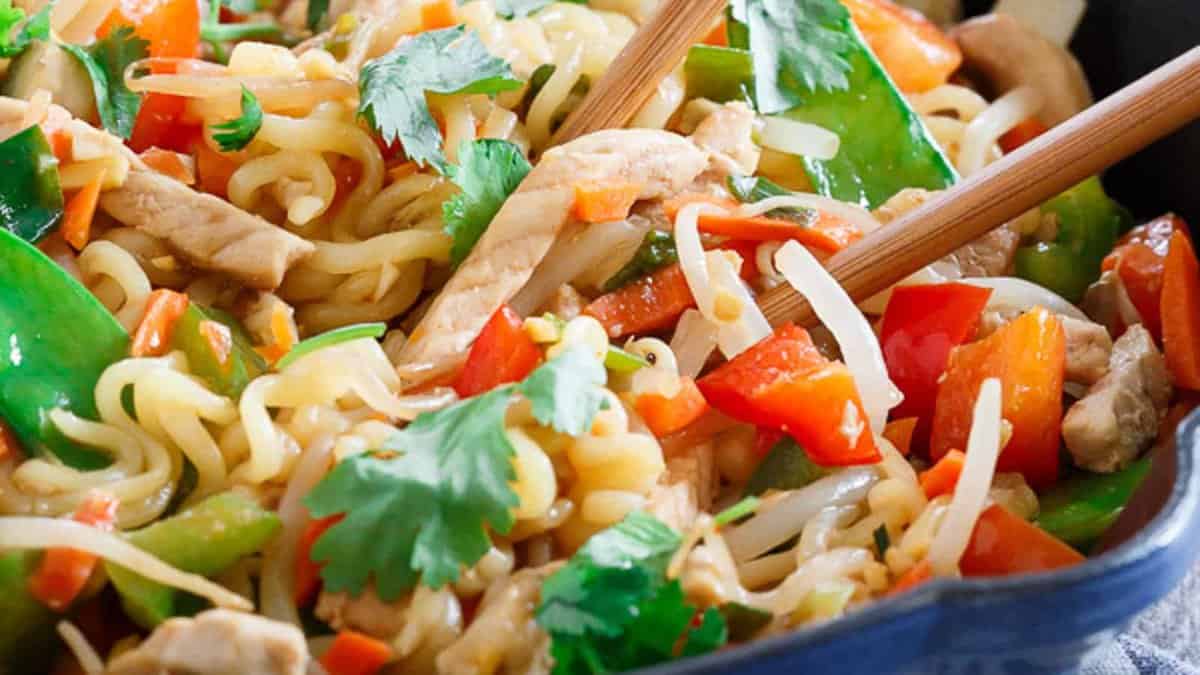 Stir-fried noodles with sliced chicken, snap peas, red bell peppers, bean sprouts, and cilantro, served with chopsticks.