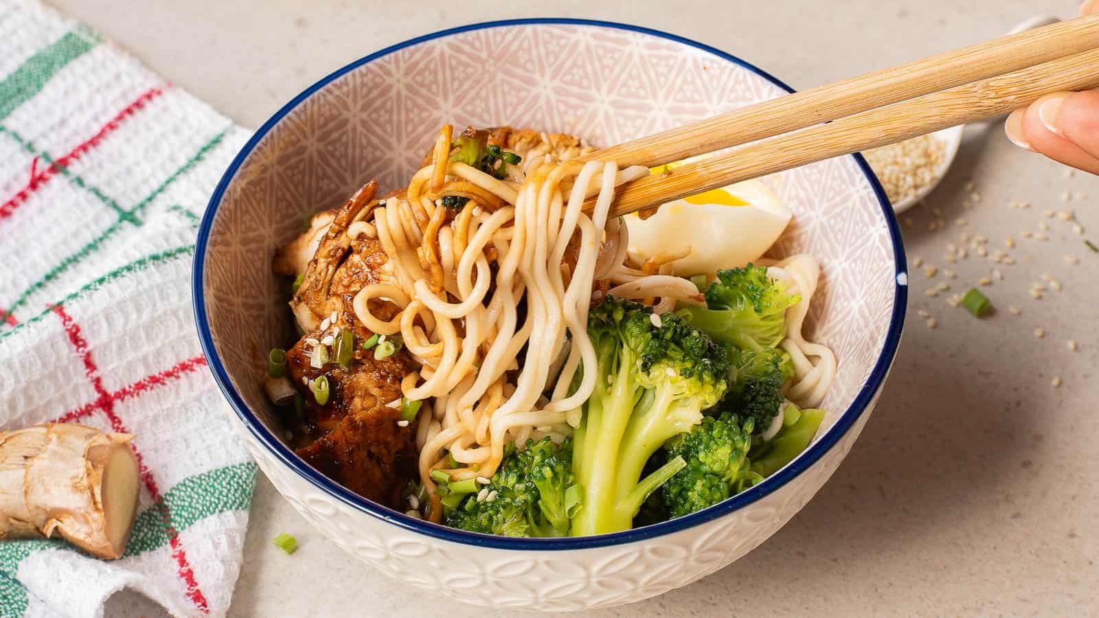 A bowl of noodles with broccoli, sliced egg, and chicken, being lifted with chopsticks, sits on a countertop next to a towel and a piece of ginger.