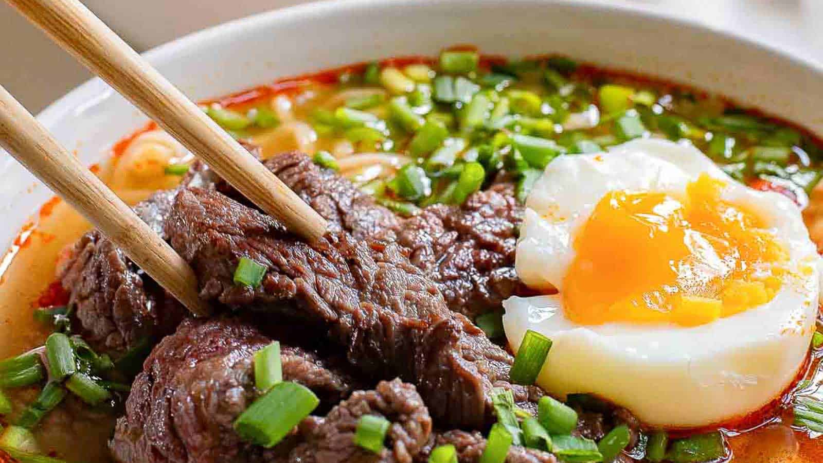 Bowl of noodle soup topped with slices of beef, chopped green onions, and a soft-boiled egg with a runny yolk; chopsticks holding a piece of beef.