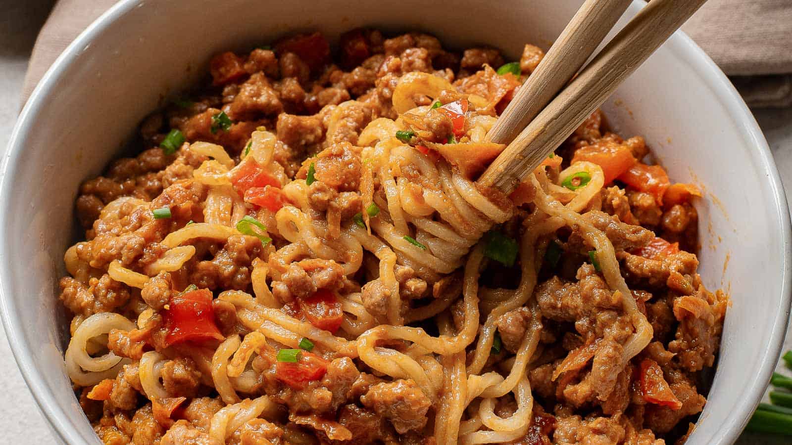 A bowl of noodles mixed with minced meat, diced red bell peppers, and chopped green onions, with chopsticks lifting a portion.