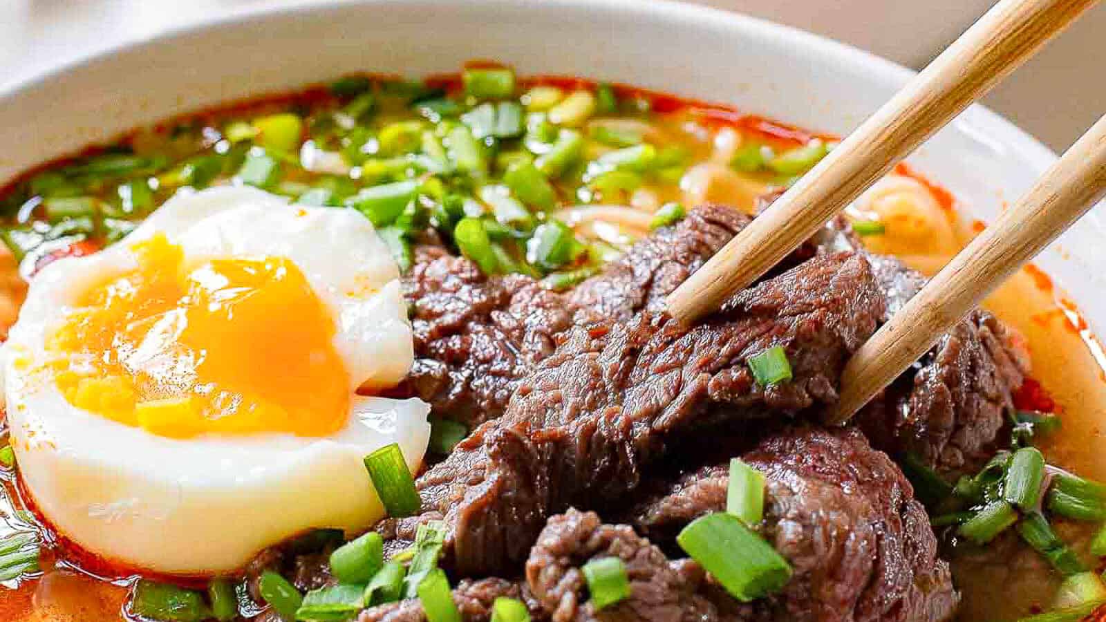 A bowl of noodle soup topped with slices of beef, a soft-boiled egg with a runny yolk, and chopped green onions, with chopsticks picking up the beef.