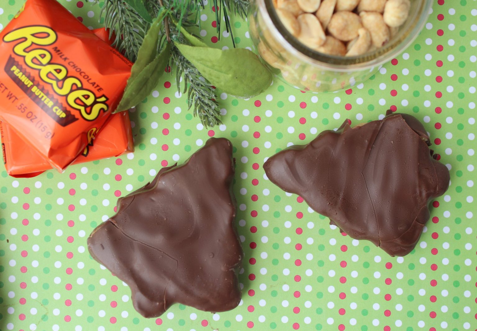 Two chocolate-covered, tree-shaped candies on a green polka dot surface, with a Reeses wrapper, peanuts in a jar, and evergreen sprigs nearby.