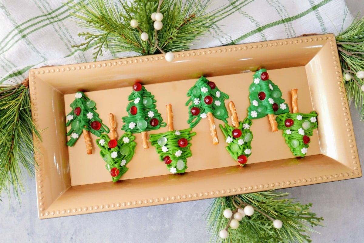 A rectangular gold tray displays Christmas tree-shaped treats decorated with green icing, red and white candies, and pretzel sticks as tree trunks, surrounded by pine branches.
