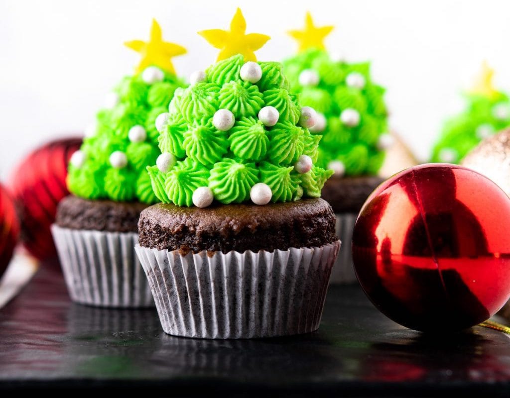 Chocolate cupcakes with green frosting decorated to look like Christmas trees, topped with yellow stars and white pearls, next to red ornaments.