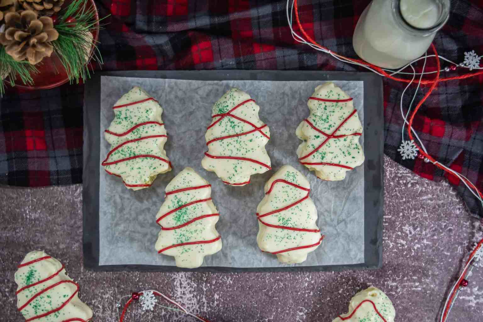Tray of Christmas tree-shaped cookies with white icing, green sprinkles, and red icing lines, placed on parchment paper; plaid cloth and a milk bottle nearby.