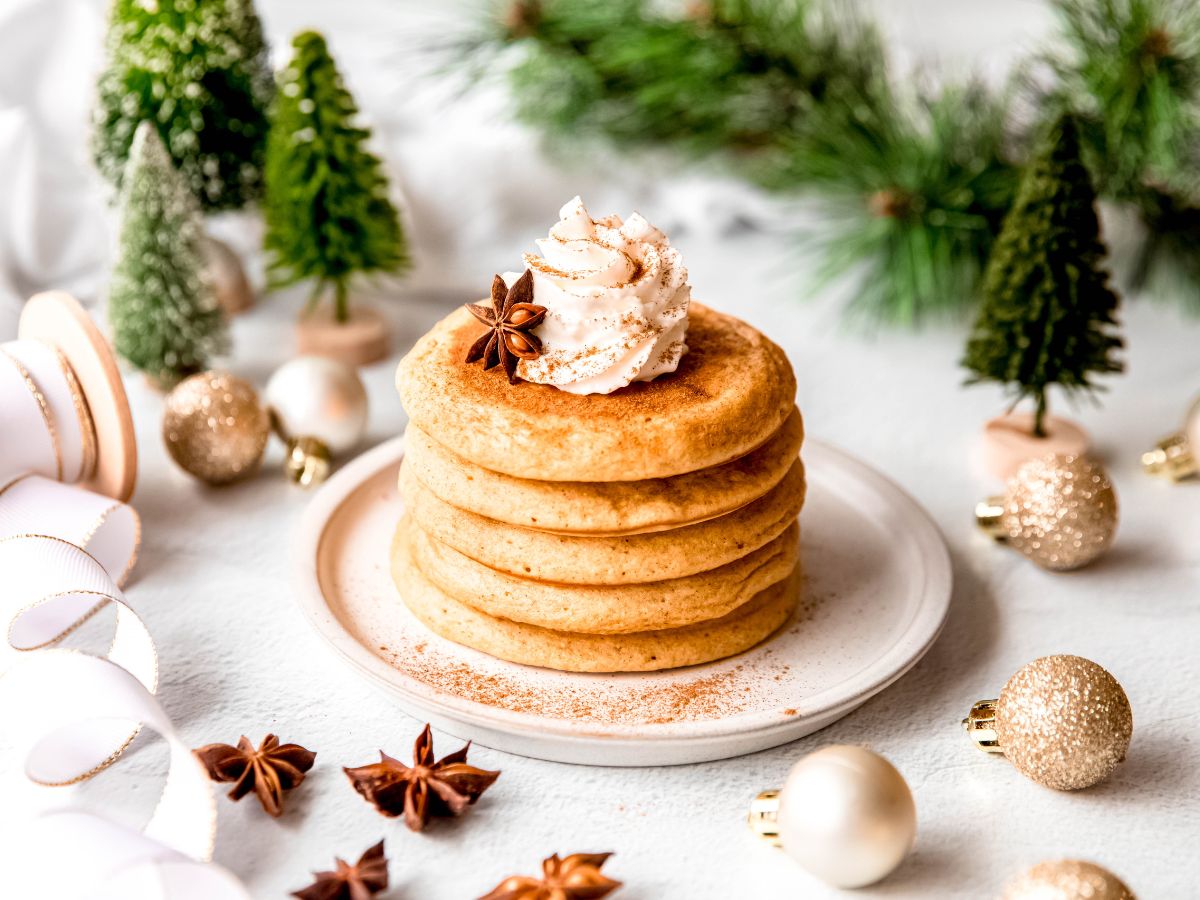 A stack of pancakes topped with whipped cream and a star anise sits on a plate, surrounded by festive decorations and small pine trees.