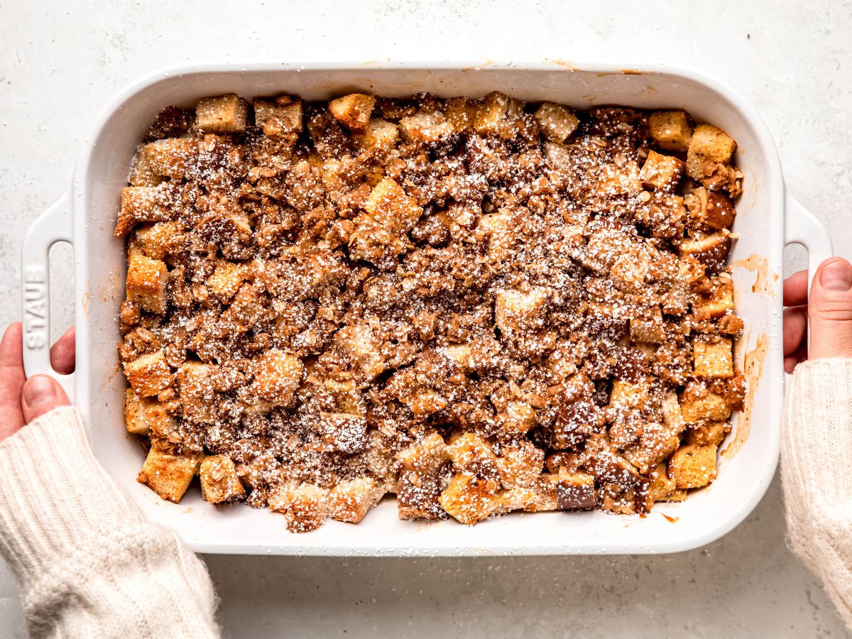A person holds a white baking dish filled with bread pudding topped with a brown crumbly mixture and powdered sugar.
