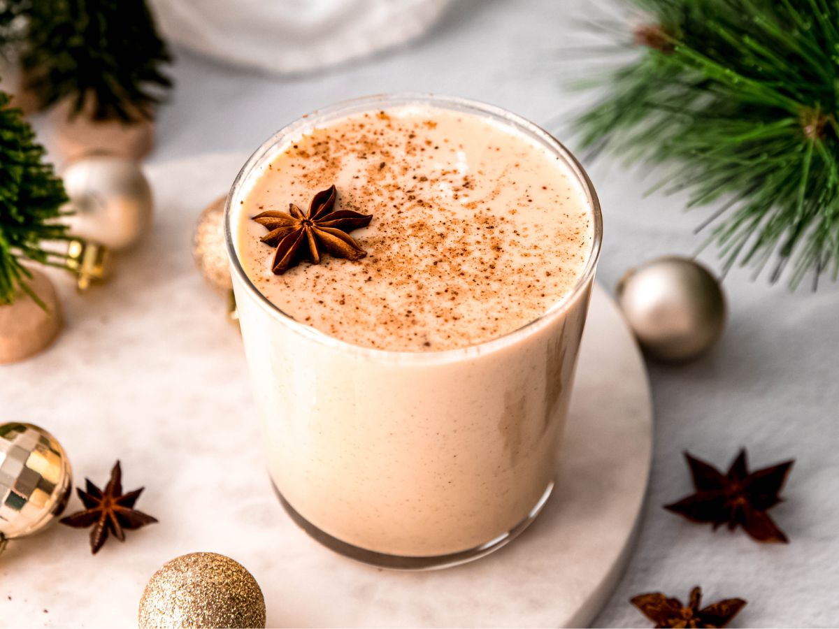 A glass of creamy eggnog topped with ground nutmeg and a star anise, surrounded by festive ornaments and pine branches.