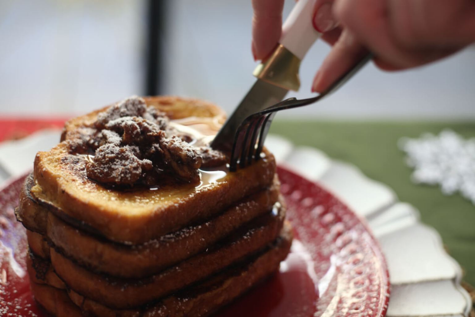 A hand uses a fork and knife to cut into a stack of French toast topped with syrup and powdered sugar on a red plate.