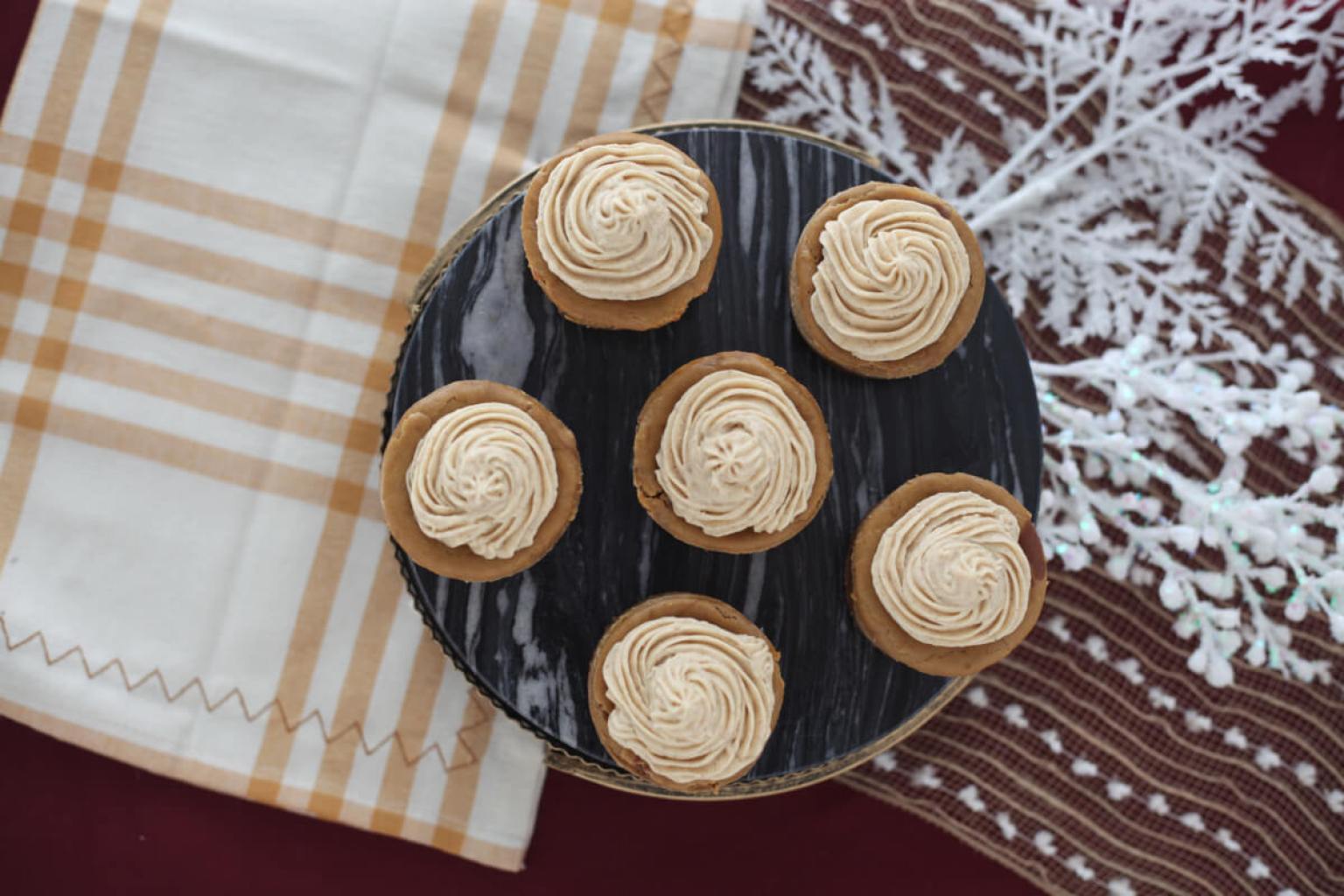 Six cupcakes with swirled frosting are arranged on a dark plate, placed on a table with a checked cloth and decorative white snowflake ornament.