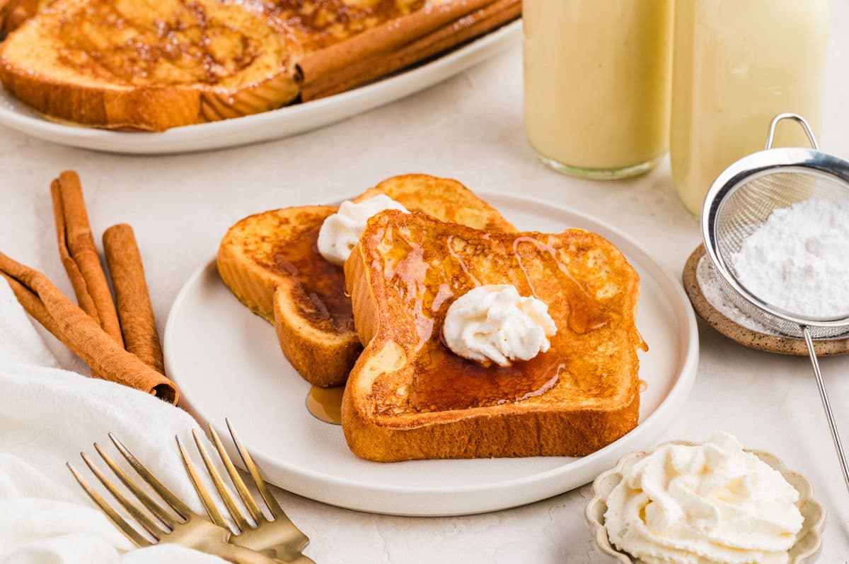 Two slices of French toast with whipped cream and syrup on a plate, surrounded by cinnamon sticks, forks, powdered sugar, and bottles of a light-colored drink.