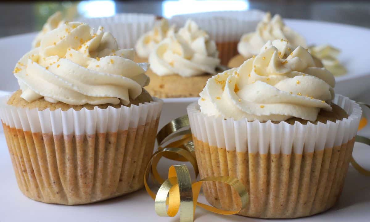 Four cupcakes with swirled white frosting sit on a white plate, decorated with gold sprinkles and gold ribbon curls.