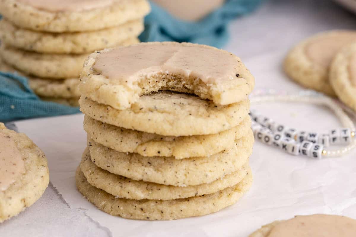 A stack of six frosted cookies, with the top cookie showing a bite taken out, sits on parchment paper. More cookies are visible in the background.