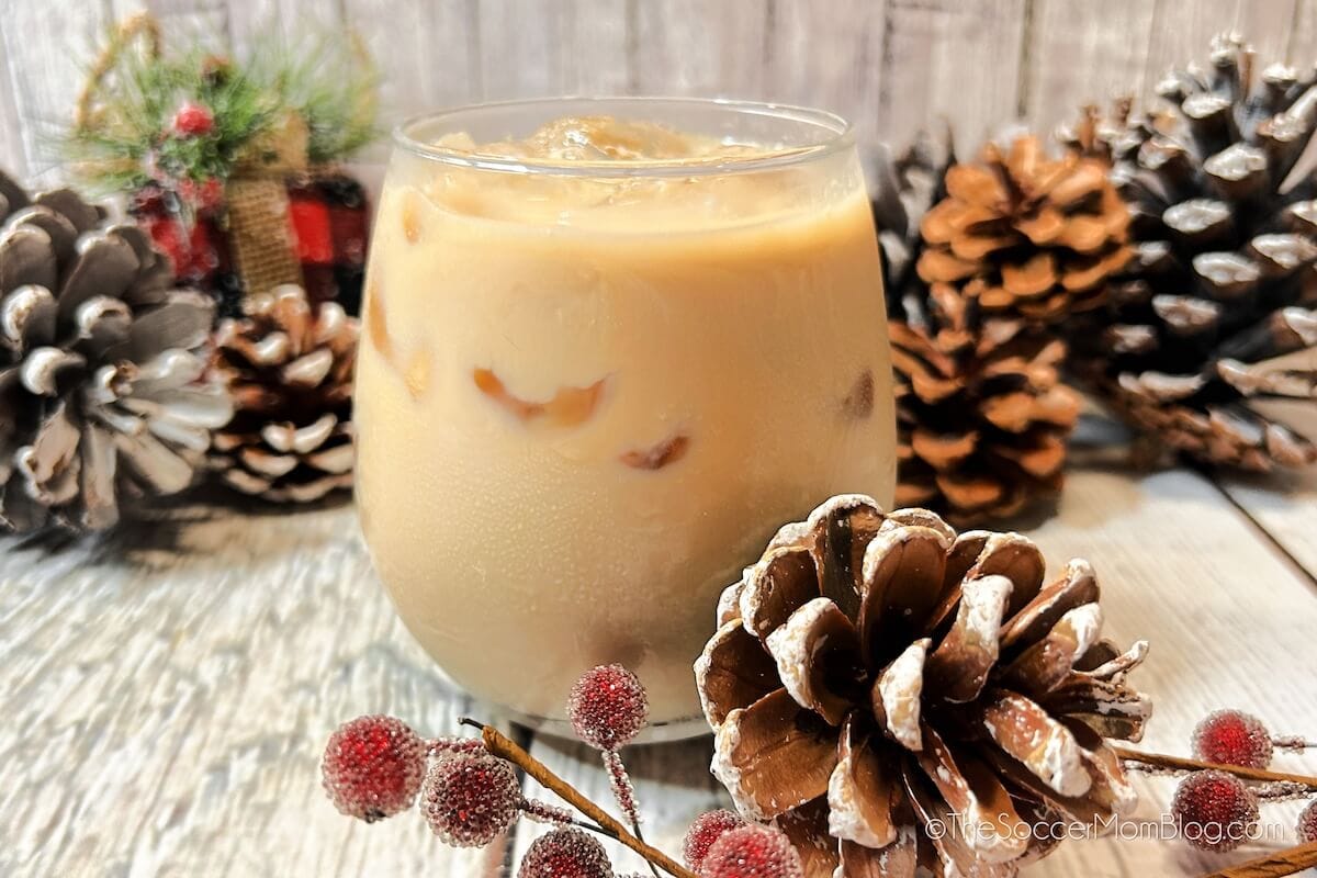 A glass of iced coffee sits on a wooden surface surrounded by pinecones, red berries, and winter-themed decor.
