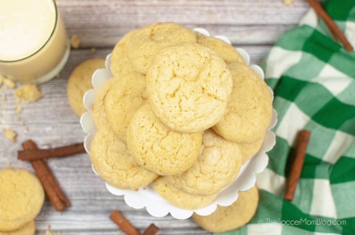 A stack of round, pale cookies on a white cake stand, surrounded by cinnamon sticks, a green checkered cloth, and a glass of milk on a wooden surface.