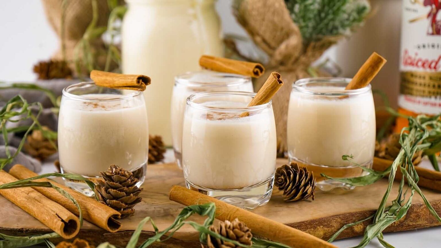 Four glasses of creamy eggnog topped with cinnamon sticks, surrounded by pinecones, cinnamon sticks, greenery, and a bottle in the background.