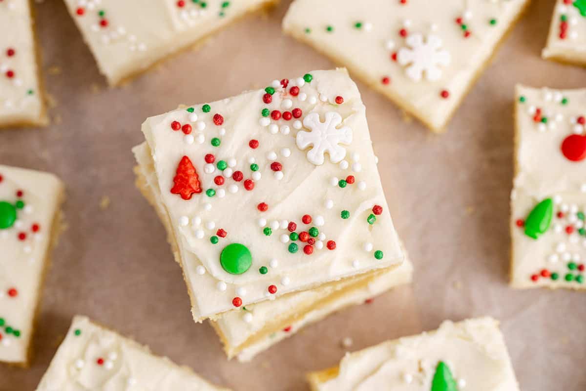 Square sugar cookie bars with white frosting, topped with red, green, and white sprinkles and holiday-themed decorations, arranged on parchment paper.