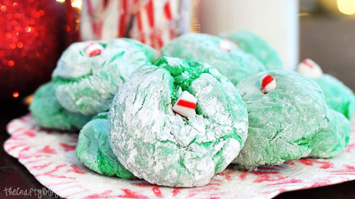 Green crinkle cookies dusted with powdered sugar, each topped with a small peppermint candy, arranged on a decorative plate.