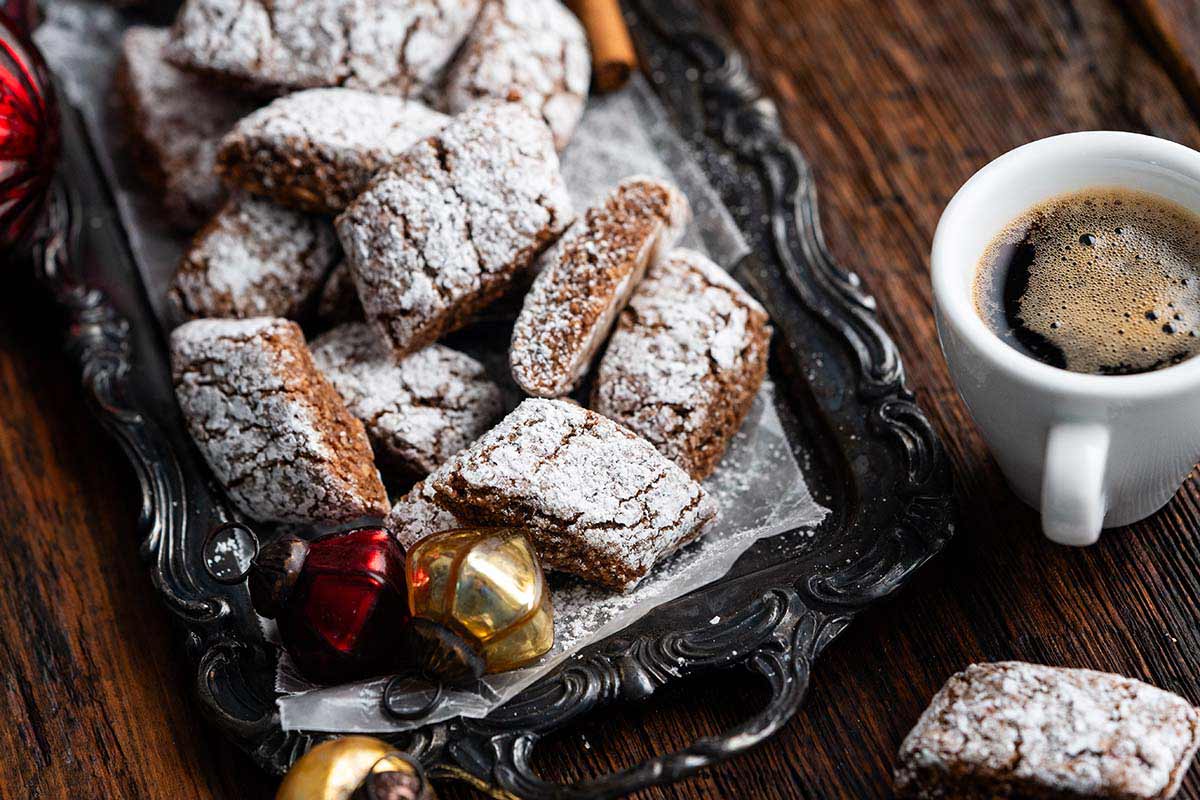 A tray of powdered sugar-dusted cookies sits next to a cup of black coffee and Christmas ornaments on a rustic wooden table.