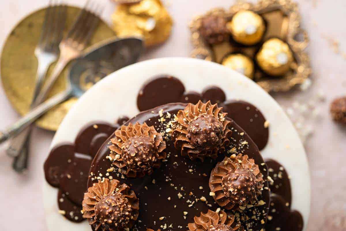 A chocolate cake topped with swirls of chocolate frosting, chopped nuts, and chocolates, set on a white plate with dessert utensils and candies nearby.