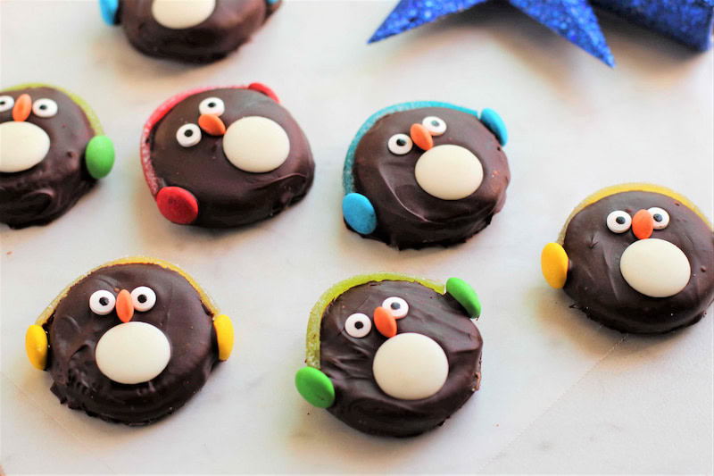 Chocolate-covered cookies decorated to look like penguin faces, with candy eyes, white candy bellies, and colorful candy earmuffs.