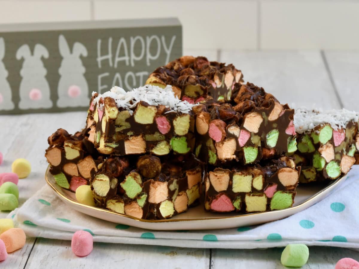 A plate of chocolate marshmallow squares with colorful mini marshmallows, set on a polka-dot napkin, with an Easter sign in the background.