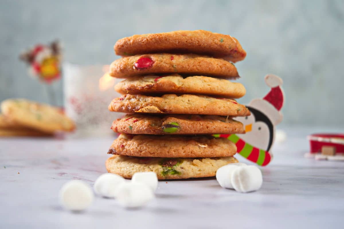 A stack of six cookies with colorful pieces sits on a marble surface, surrounded by mini marshmallows and holiday-themed decorations.
