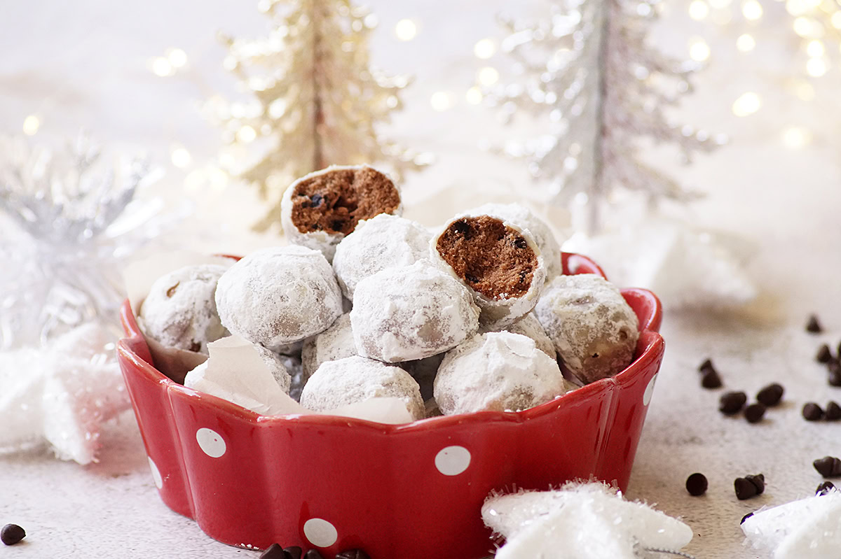 A red polka dot bowl filled with powdered sugar-coated chocolate balls, some with a bite taken out, sits in front of white decorative trees and lights.