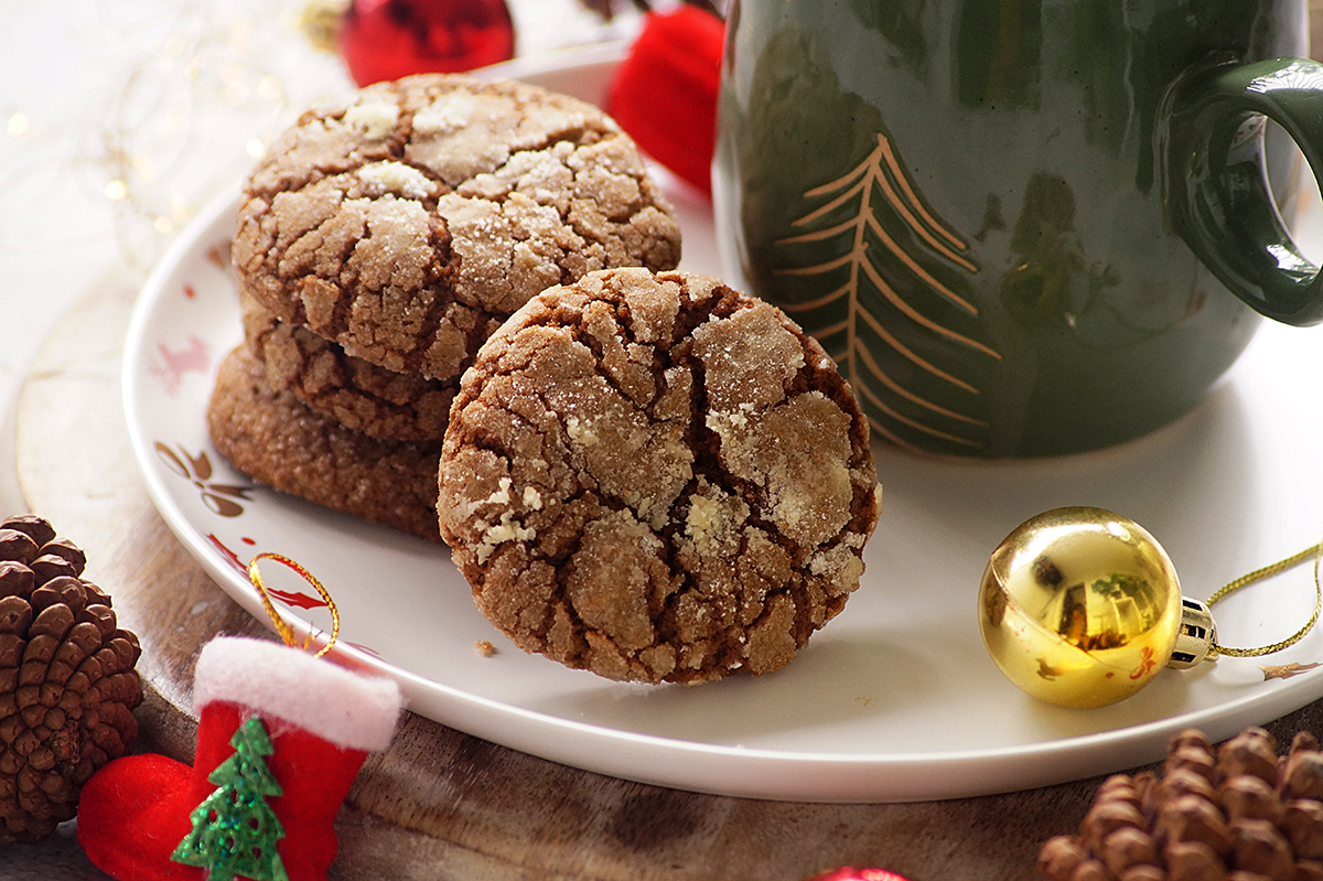 A plate with three crinkle cookies beside a green mug with a tree design, surrounded by pinecones, ornaments, and festive holiday decorations.