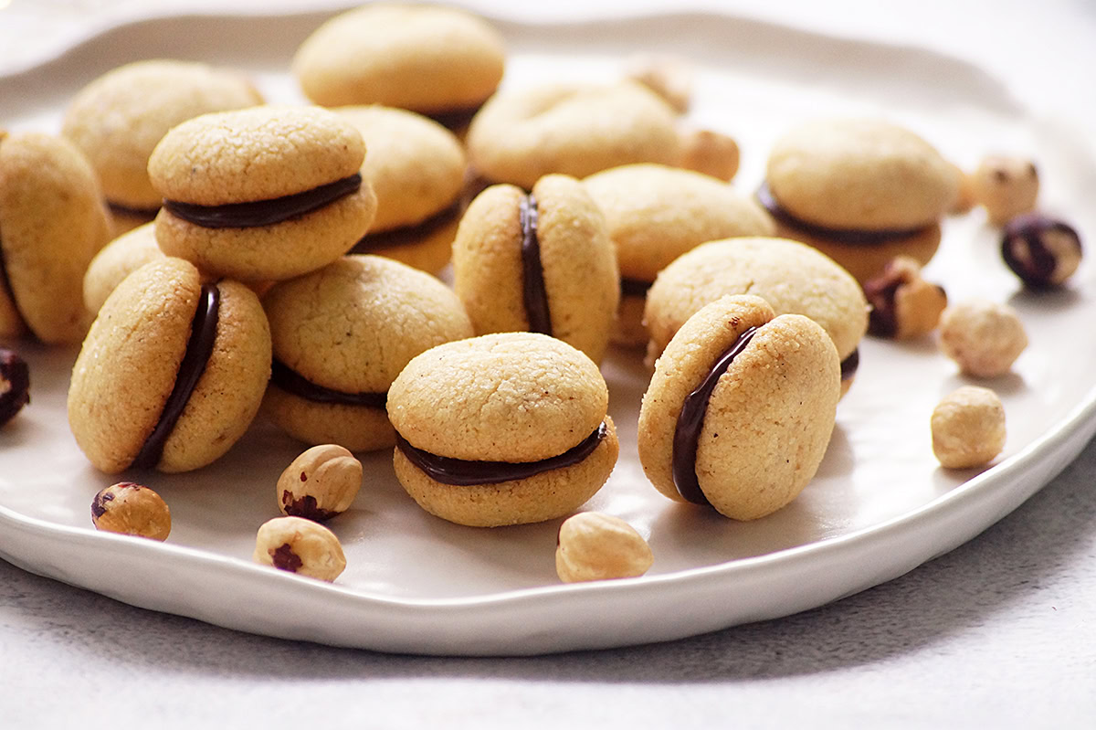 A plate of sandwich cookies with chocolate filling is arranged with scattered hazelnuts on a white surface.