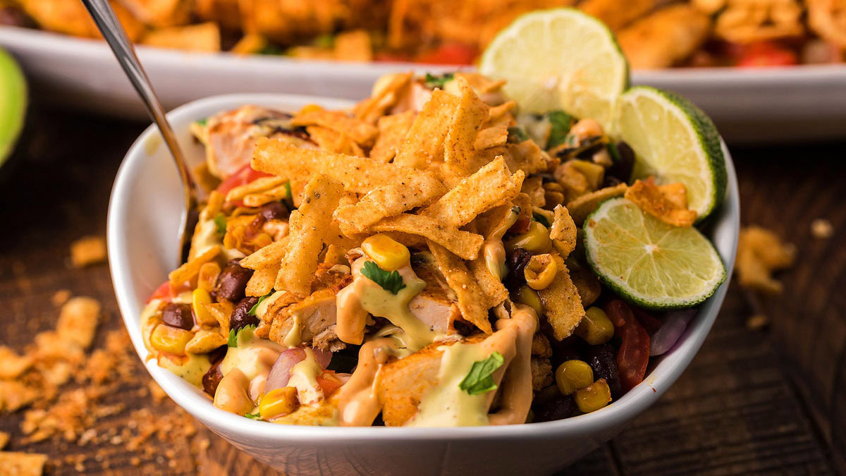 A bowl of southwestern salad topped with corn, beans, tortilla strips, creamy dressing, and lime slices, with a fork on the side.