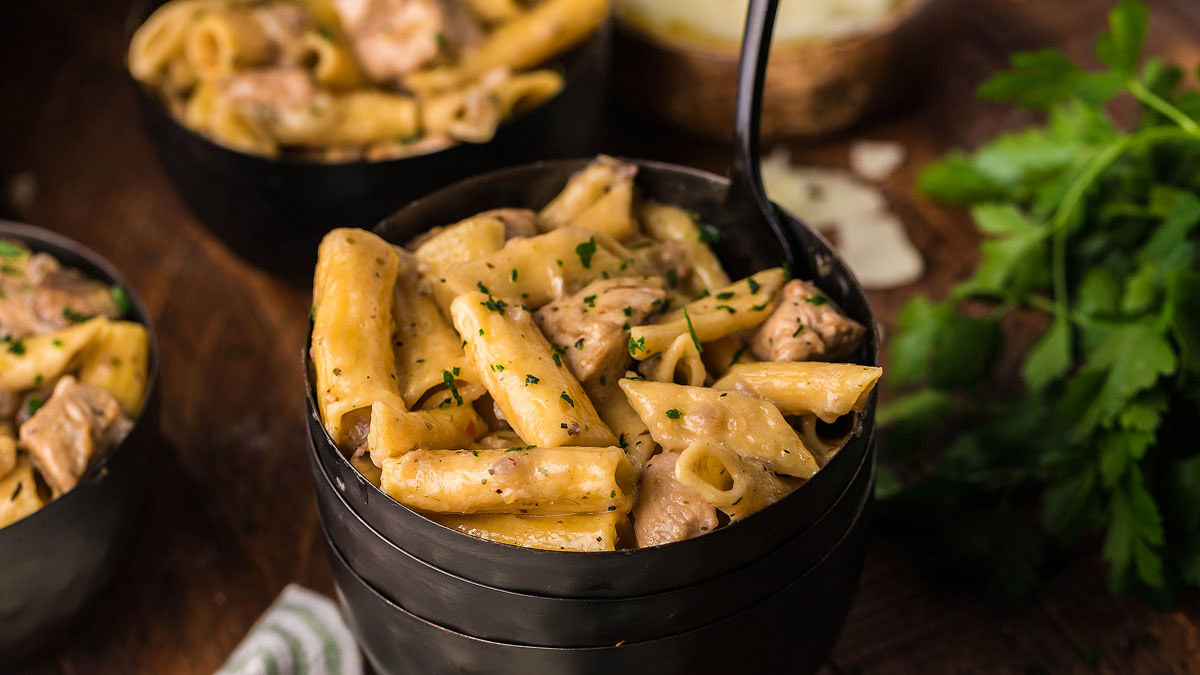 A black bowl filled with creamy pasta and chunks of chicken, garnished with herbs, sits on a wooden table next to fresh parsley.