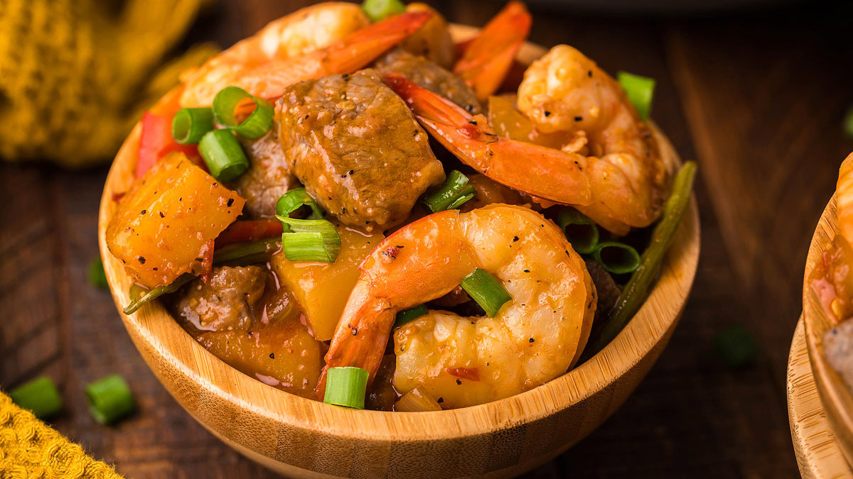 A wooden bowl filled with shrimp, beef chunks, pineapple pieces, and chopped green onions in a savory sauce.