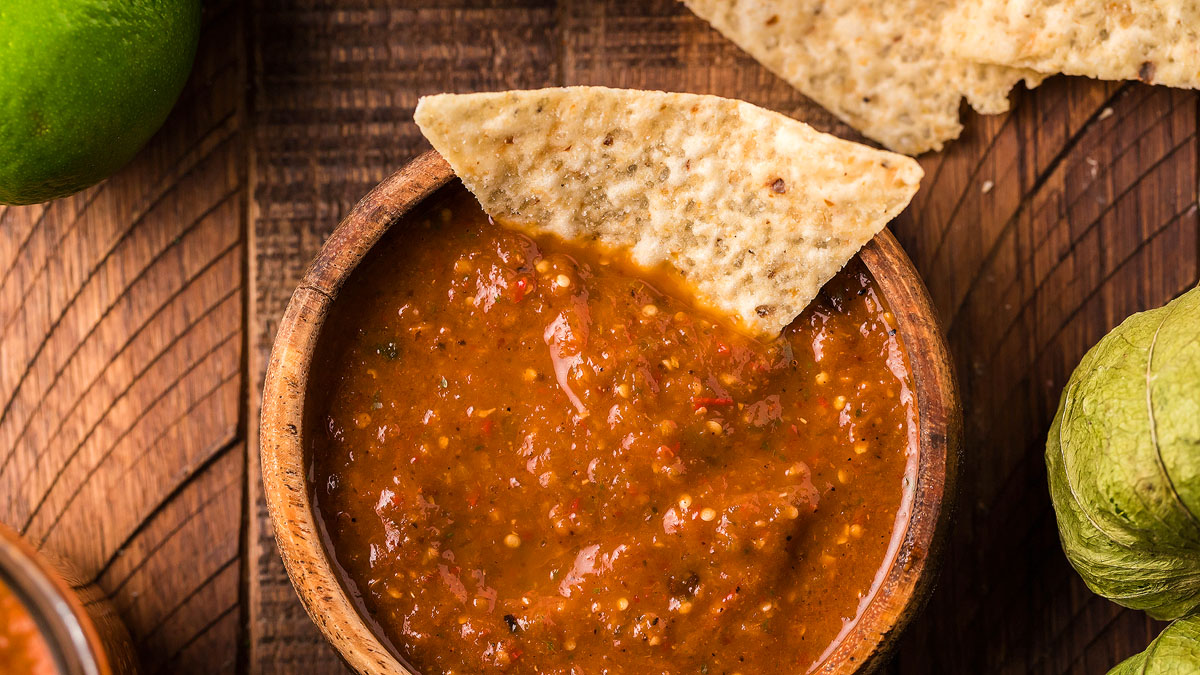 A tortilla chip dipped into a bowl of red salsa, with more chips and a lime visible on a wooden surface.