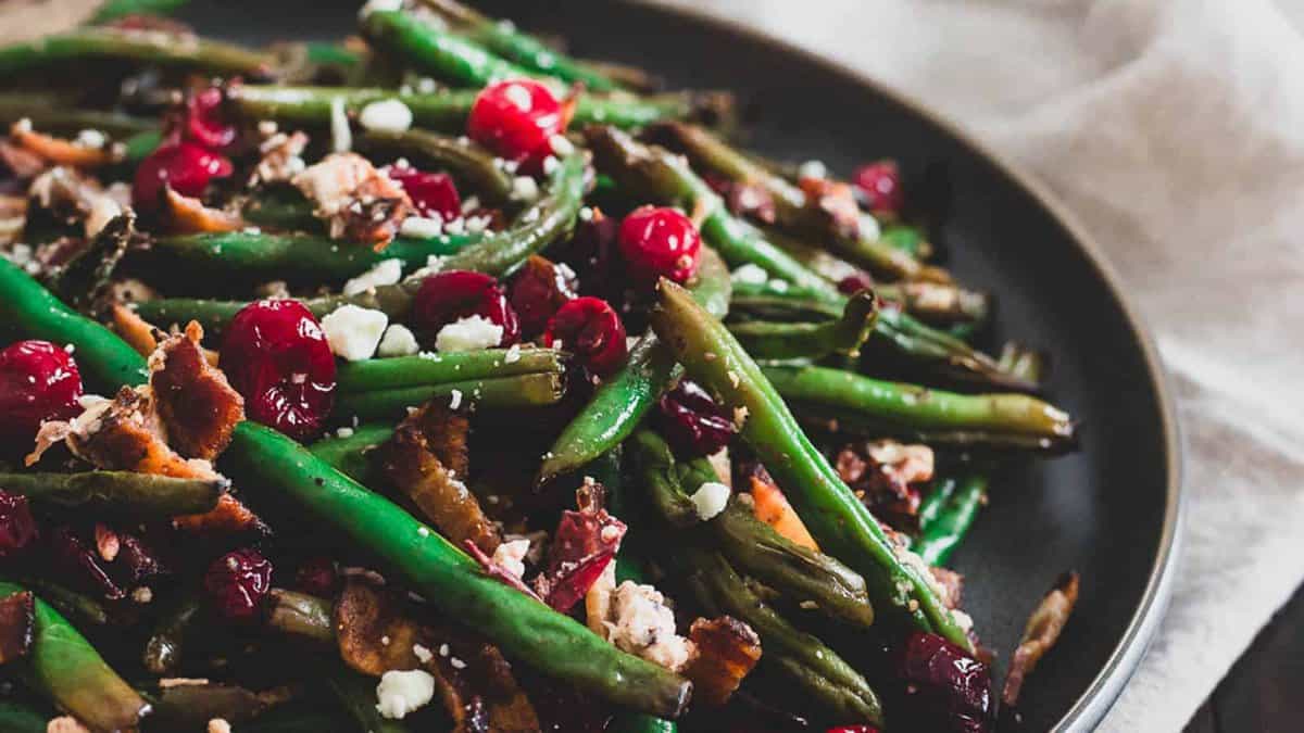 A plate of roasted green beans with bacon, dried cranberries, and crumbled cheese, served on a dark dish.