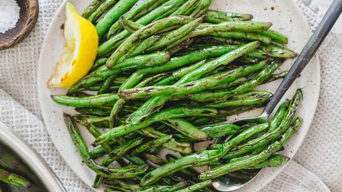 A plate of grilled green beans with a lemon wedge and a serving spoon on the side.