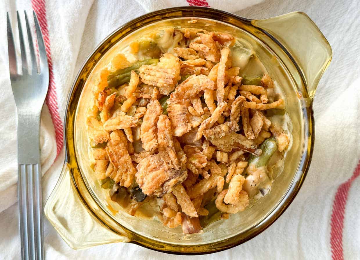 A glass dish filled with green bean casserole topped with crispy fried onions, placed on a white cloth with a fork beside it.