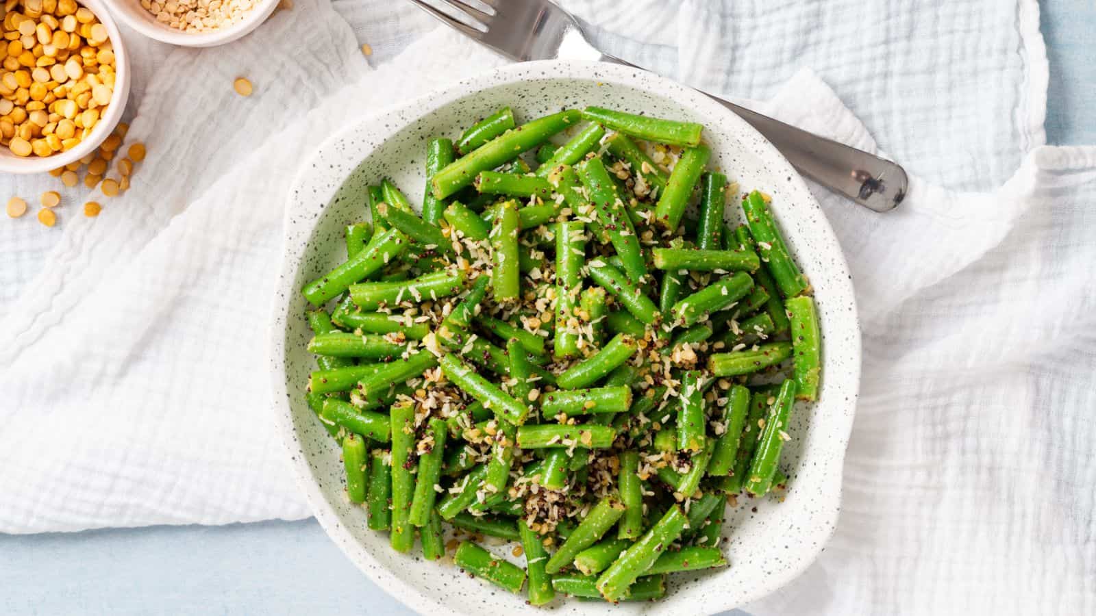 A white bowl filled with cooked green beans topped with sesame seeds, placed on a white cloth with a fork and small bowls of ingredients nearby.